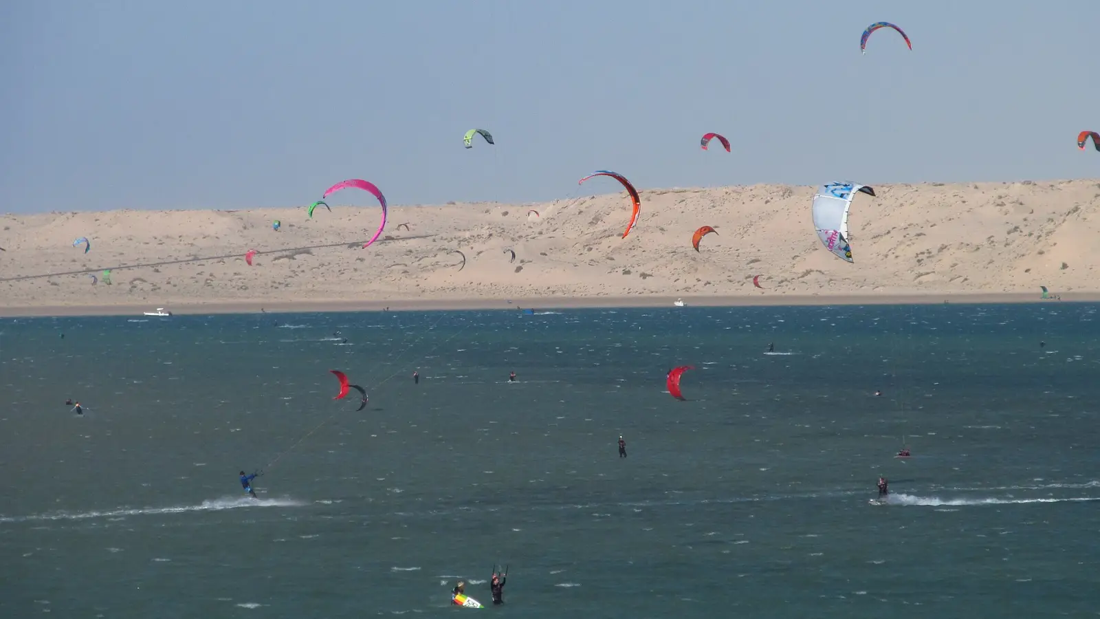 Tientallen kites boven een vlakke lagune, met een zandduin erachter