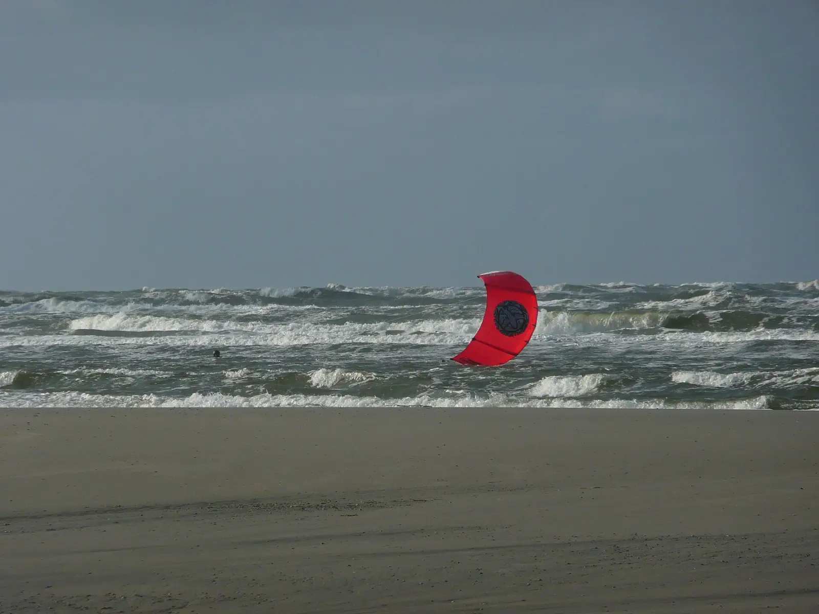Een rode kite ligt neergestort in de branding van de Noordzee onder een donkere lucht