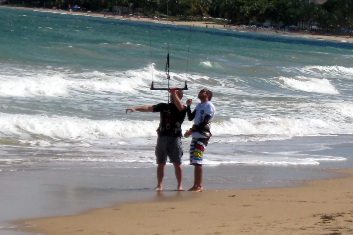 An instructor stands on the beach right next to a student holding the bar of a kite